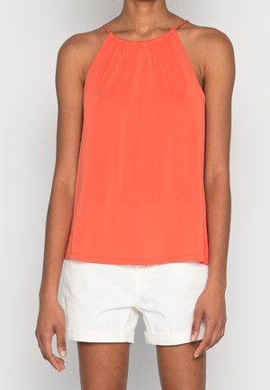 Frau trägt ärmelloses orangefarbenes Neckholder-Top und weiße Shorts mit umgeschlagenem Saum, steht vor einem schlichten hellen Hintergrund.