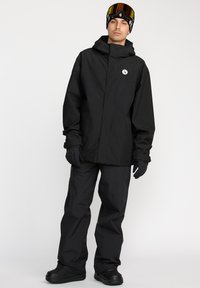 Veste d'hiver noire et pantalon en matériau imperméable, avec une capuche, des poignets élastiques et un patch logo, portés avec des gants noirs et des lunettes de protection.