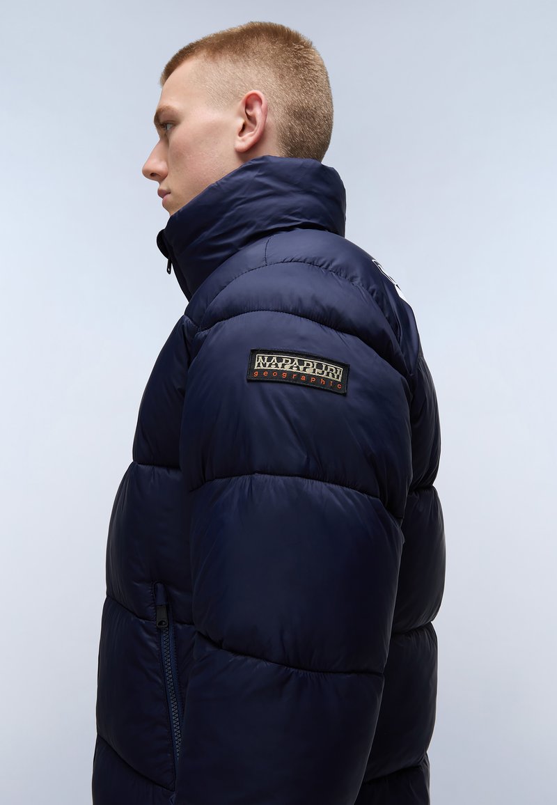 Granatowa kurtka puchowa z wysokim kołnierzem, pikowanym wzorem, boczną kieszenią na zamek oraz naszywką z napisem "Napapijri Geographic" na rękawie.