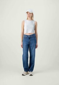 Fehér bordázott crop top, magasan derekú kék farmernadrág és világos sportcipők. A modellen fehér sapka van. Egyszerű háttér.