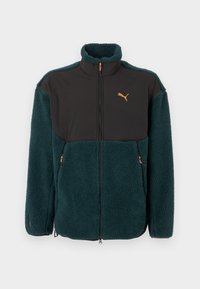 Sinivihreä fleecetakki mustilla kangaspaneeleilla, korkealla kauluksella, edessä vetoketjusuljenta, vetoketjulliset taskut ja oranssi logodetalji. Pehmeä rakenne.