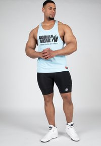 Lichtblauw sporttopje met zwart "GORILLA WEAR" logo, zwarte compressiekorte broek, en witte sportchoenen met logo. Eenvoudig en functioneel ontwerp.