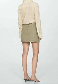 Femme portant une chemise beige à manches longues rentrée dans une mini-jupe verte olive avec fermeture éclair au dos, associée à des sandales à talons couleur nude.