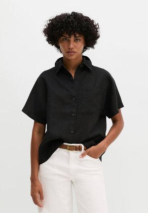 Vrouw met krullend haar, die een losse zwarte korte mouwen blouse met knopen en een witte broek met een bruine riem draagt, staand met één hand in de zak.