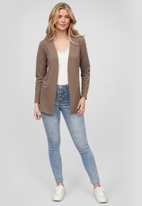 Cardigan long marron avec un tissu texturé, devant ouvert et deux poches zippées. Associé à un débardeur en dentelle blanche et un jean skinny bleu.