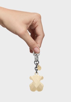 Ręka trzyma srebrno-czarny brelok z fasetowanym złotym misiem oraz małą złotą zawieszką z grawerem „TOUS”.