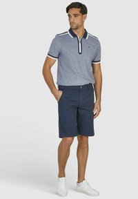 Graues Poloshirt mit weißen Akzenten, kombiniert mit marineblauen Shorts. Das Modell steht in weißen Sneakers und präsentiert eine lässige Outdoor-Bekleidung.