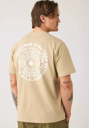 Hombre con cabello castaño que lleva una camiseta beige con texto blanco circular en la espalda, de pie de lado con las manos en los bolsillos contra un fondo liso.