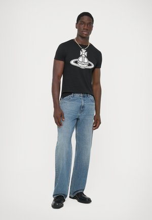 Man wearing black graphic t-shirt, light blue straight-leg jeans, black shoes, and a beaded necklace, standing against a white background.