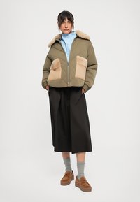Donna che indossa una giacca imbottita verde oliva con tasche e colletto in pelliccia beige, pantaloni larghi neri, calze grigie e scarpe con piattaforma marroni.