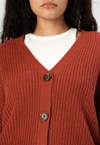 Rustfarget strikket cardigan med V-hals, prydet med to mørke knapper og ribbestruktur. Laget over en hvit ribbestrikket topp.