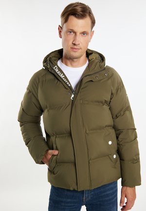 Mann mit kurzen braunen Haaren, der eine olivgrüne Daunenjacke über einem weißen Hemd und blauen Jeans trägt, Hand in der Jackentasche.