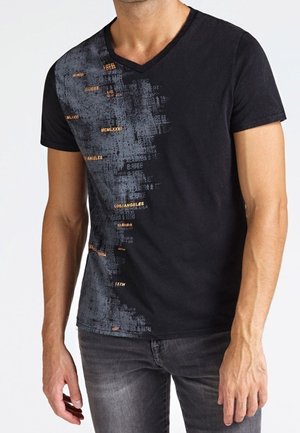 Mężczyzna w dopasowanej czarnej koszulce z dekoltem w serek, ozdobionej abstrakcyjnym szaro-pomarańczowym nadrukiem, w połączeniu z ciemnoszarymi jeansami.