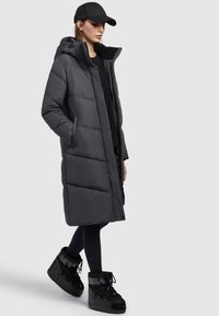Long black puffer coat with a hood, featuring quilted sections and large side pockets. Worn with black leggings and high-ankle boots.