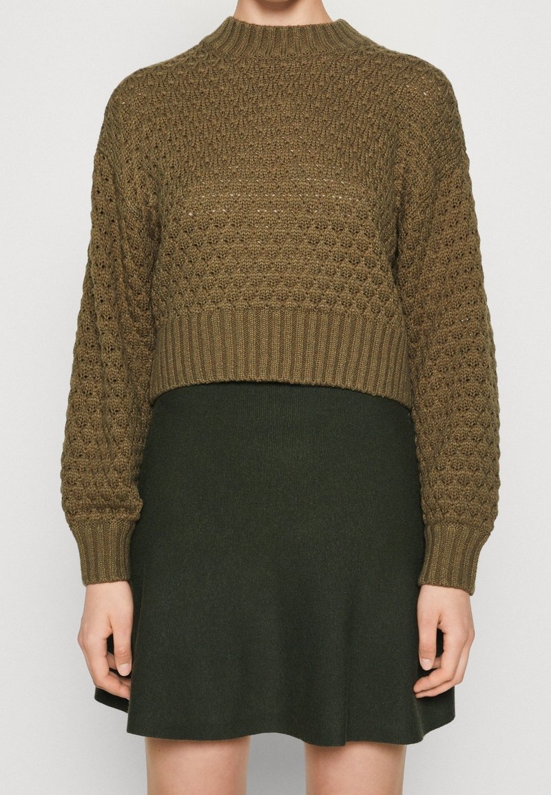 Personne portant un pull court tricoté vert olive avec un motif texturé et une jupe trapèze vert foncé.