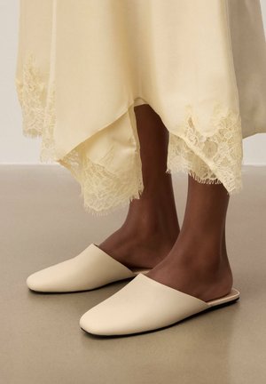 Persoană purtând pantofi crem slip-on cu talpă plată și o fustă crem cu dantelă, stând pe un podea netedă bej.