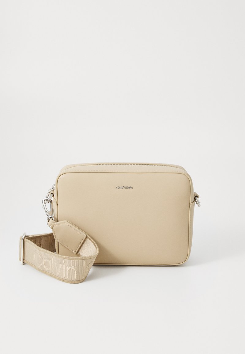 Beige rechteckige Umhängetasche aus glattem synthetischen Material, mit einem strukturierten Gurt und silberfarbenen Hardware-Elementen. Logo auf der Vorderseite angebracht.