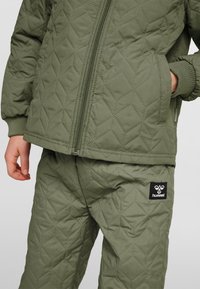 Dziecko ubrane w oliwkowo zieloną pikowaną kurtkę i spodnie z zamkiem błyskawicznym oraz kieszeniami, widoczne logo hummel na spodniach.