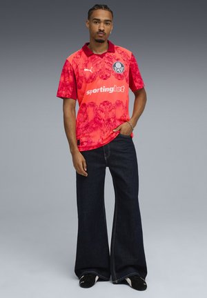 Rød blomster mønstret sportstrøje med krave og logo, parret med brede mørkeblå denim jeans og sorte fodtøj.