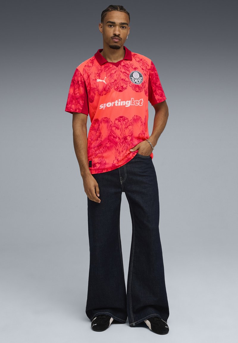 Jersey deportivo con un patrón floral rojo que presenta un cuello y un logo, combinado con unos pantalones vaqueros de pierna ancha en denim oscuro y calzado negro.