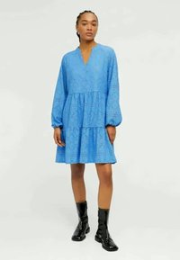 Robe bleue à volants en tissu en dentelle. Dotée de longues manches bouffantes et d'un décolleté en V. Porte des bottines noires brillantes.