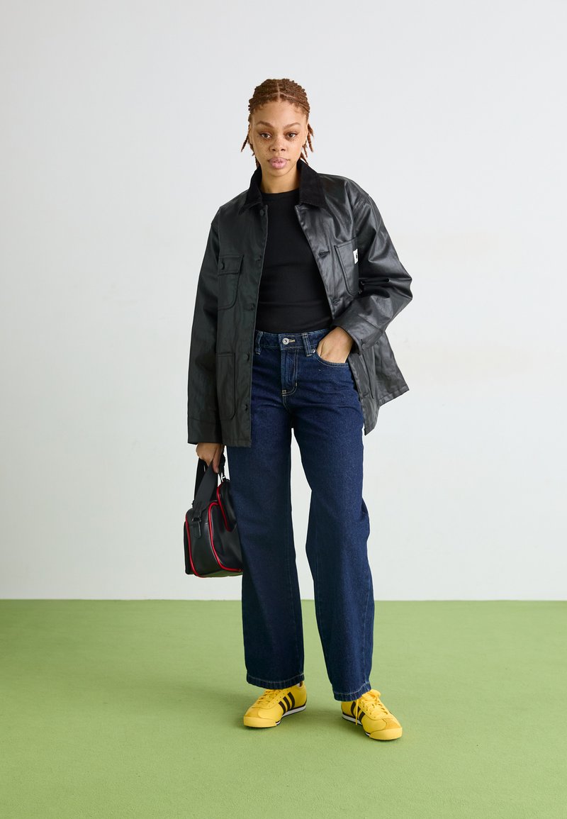 Giacca di pelle nera con un design colletto, abbinata a jeans a gamba larga blu scuro e sneakers gialle. Il modello tiene una borsa nera con accenti rossi.