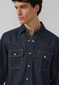 Donker spijkerblouse met een knoopsluiting aan de voorkant, twee borstzakken met kleppen en contrasterende gele stiksels. Klassiek kraagontwerp.