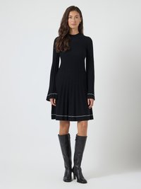 Mulher com cabelo longo e ondulado, vestindo um vestido pregueado preto com acabamento branco e botas de couro pretas até o joelho, em pé contra um fundo branco liso.