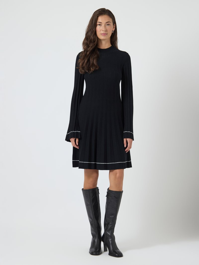 Mulher com cabelo longo e ondulado, vestindo um vestido pregueado preto com acabamento branco e botas de couro pretas até o joelho, em pé contra um fundo branco liso.