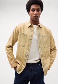 Cămașă button-up de un bej deschis, fabricată din denim moale, având două buzunare la piept, guler și o croială relaxată. Texturată cu o ușoară decolorare.