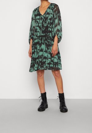 Vrouw draagt een loszittende groene en zwarte jurk met een patroon tot op de knie, met lange mouwen en zwarte veterlaarzen, staand op een grijze vloer.