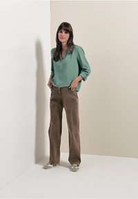 Türkise Bluse mit einem Knopfdekolleté, kombiniert mit braunen weiten Hosen. Weiche Texturen, klare Linien und lässiges Schuhwerk runden den Look ab.