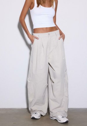 Žena nosi široké béžové nohavice, biely krátky tielko a biele tenisky, stojí s rukami v vreckách pred jednofarebným pozadím.