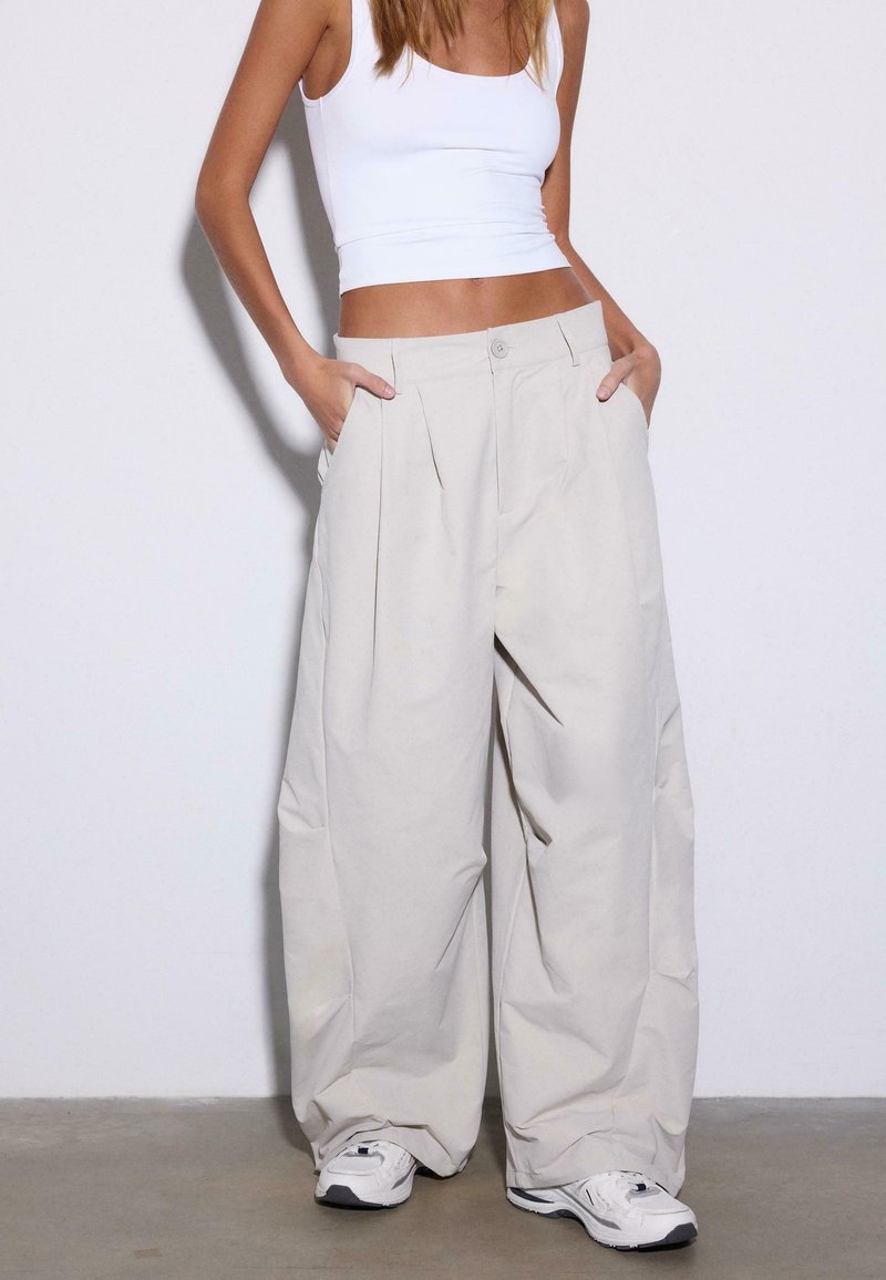Femme portant un pantalon beige à jambes larges, un débardeur court blanc et des baskets blanches, debout les mains dans les poches devant un fond uni.