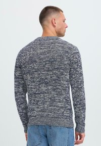 Man som vänder bort mot kameran, iklädd en marinblå och vit melerad stickad tröja samt ljusblå jeans, mot en vit bakgrund.