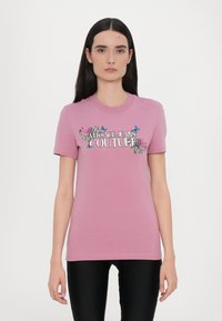Rosa bomulls t-skjorte med korte ermer, med et blomsterdesign og teksten "VERSACE JEANS COUTURE" i fet skrift.