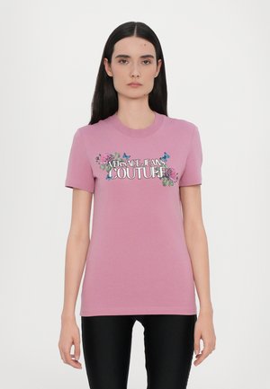 T-shirt in cotone rosa con maniche corte, caratterizzata da una grafica floreale e la scritta "VERSACE JEANS COUTURE" in caratteri in grassetto.