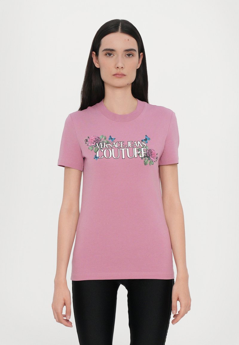 Rosa bomulls t-skjorte med korte ermer, med et blomsterdesign og teksten "VERSACE JEANS COUTURE" i fet skrift.