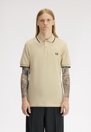 Jeune personne aux longs cheveux blonds et tatouages couvrant tout le bras, portant un polo beige avec bordure noire et un pantalon noir, debout devant un fond blanc.