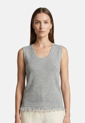 Donna con capelli castani lisci che indossa un top grigio senza maniche in maglia con orlo sfrangiato e pantaloni beige chiari, in piedi su sfondo bianco.