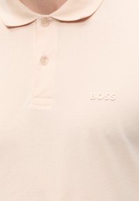Polo rose clair avec deux boutons et logo "BOSS" en relief sur la poitrine gauche, porté par une personne visible du cou au torse.