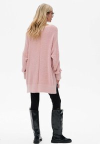 Roze gebreide cardigan met een losse pasvorm, geribbelde zomen en lange mouwen, gecombineerd met zwarte aangesloten broek en kniehoge zwarte laarzen.