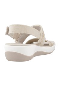 Sandalia beige con tiras de tejido trenzado y una suela blanca acolchada. El diseño incluye un soporte trasero y un agarre texturizado en la suela exterior.