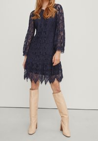Robe en dentelle navy à manches longues, motif floral et ourlet festonné. Portée avec des bottes beiges hautes jusqu'aux genoux avec un talon texturé.