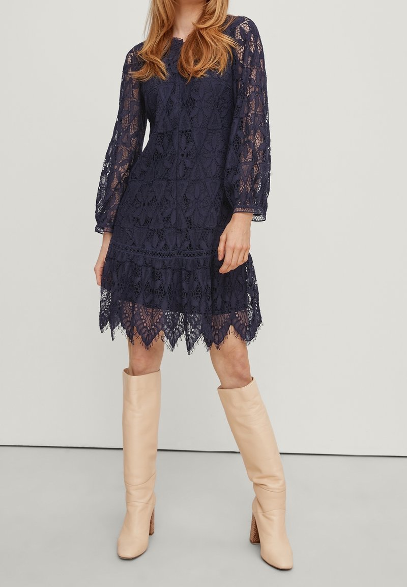 Robe en dentelle navy à manches longues, motif floral et ourlet festonné. Portée avec des bottes beiges hautes jusqu'aux genoux avec un talon texturé.