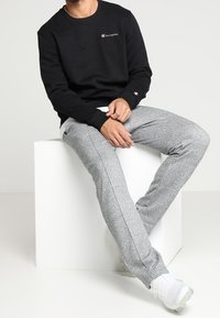 Felpa nera con piccolo logo ricamato, abbinata a pantaloni della tuta grigi chiari a sigaretta, con tessuto a trama e strisce laterali. Scarpe bianche.