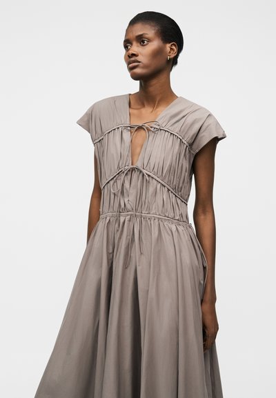 Žena s krátkymi vlasmi oblečená v voľných taupe šatách so zdobením a stiahnutými šnúrkami na hrudníku a v páse, stojaca pred jednoduchým pozadím.