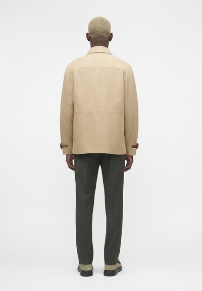 Άτομο με κοντά ξανθά μαλλιά στέκεται γυρισμένο με την πλάτη φοράει μια μπεζ πανωφόρι, σκούρο παντελόνι και μπεζ παπούτσια σε απλό λευκό φόντο.