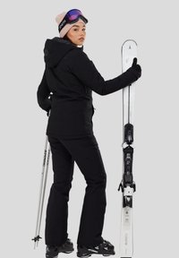 Vrouw in zwart skipak die ski's en stokken vasthoudt, draagt een roze muts en paarse skibril, staand tegen een grijze achtergrond.