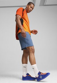 Męska odzież sportowa: pomarańczowa i czarna koszulka z krótkim rękawem, niebieskie szorty, niebieskie sneakersy z pomarańczowymi sznurówkami oraz białe skarpetki z czarnym logo.